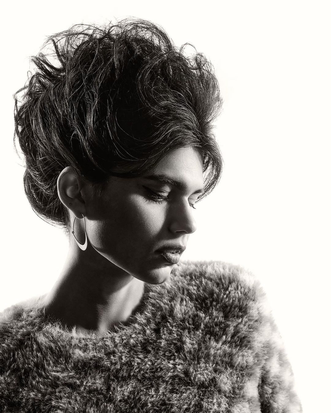 Monochrome portrait of a person with dark hair in an updo wearing hoop earrings and a textured fur-like top against a white backdrop.
